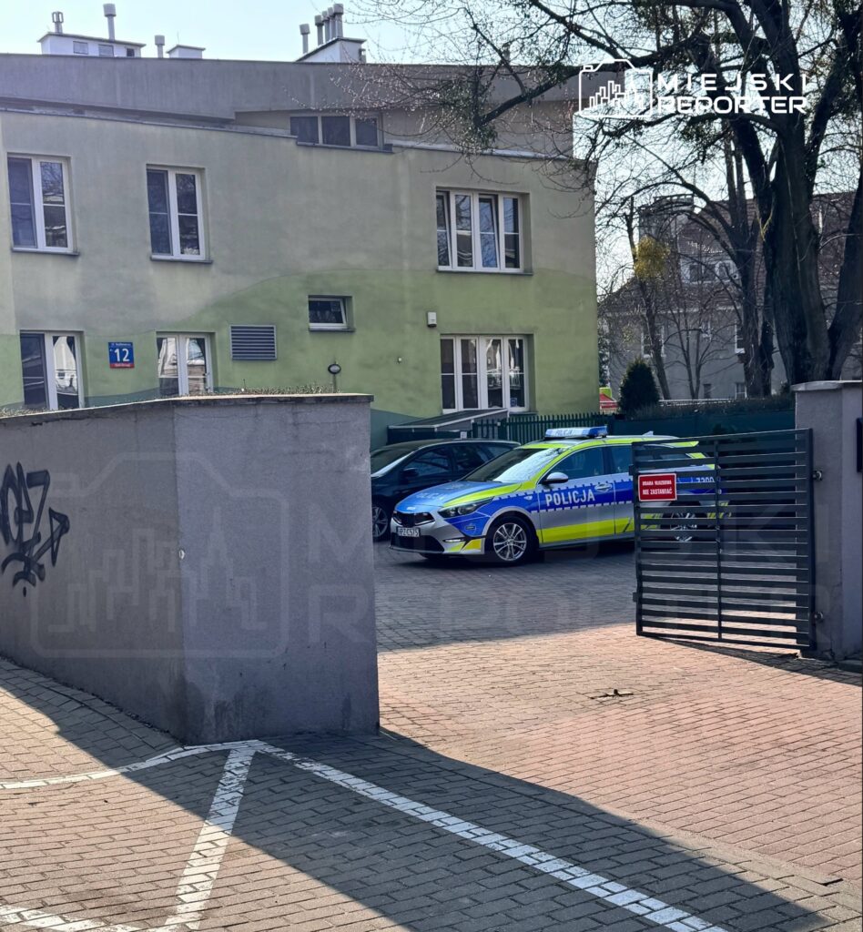 Policyjny radiowóz z oznaczeniem "POLICJA" zaparkowany przed budynkiem z numerem 12, otoczony zielenią.