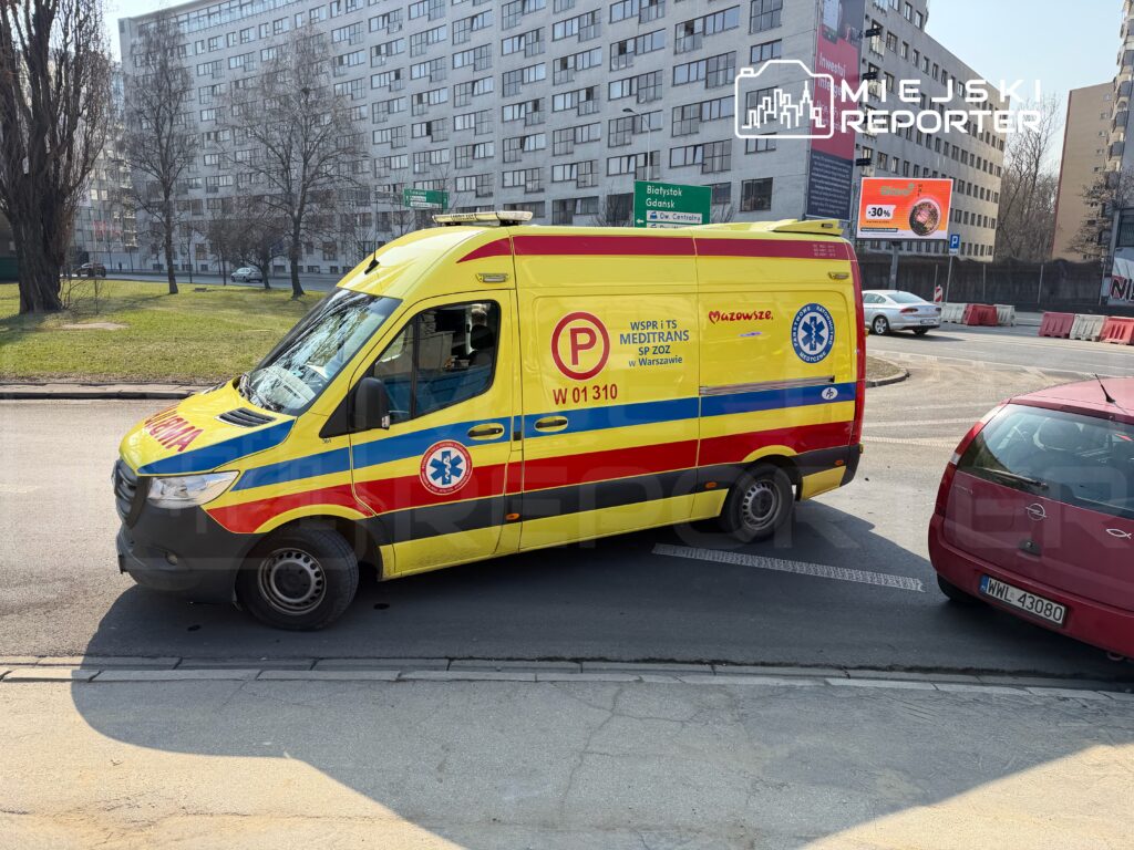 Karetka pogotowia w kolorach żółto-czerwonych zatrzymana na skrzyżowaniu w miejskim otoczeniu.