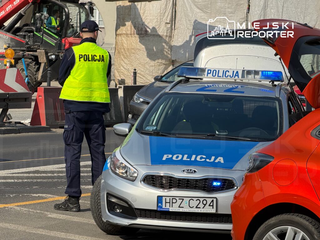 Policjant w kamizelce odblaskowej kontroluje ruch przy policyjnym samochodzie na zbudowanej drodze.