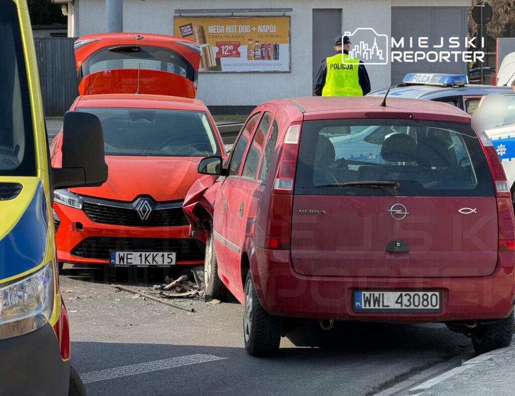 Dwa uszkodzone samochody, czerwony Opel Meriva i pomarańczowy Renault, zderzyły się na ulicy, obok policjanta w kamizelce.