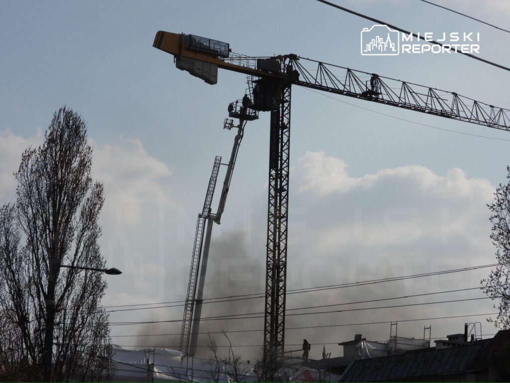 Dźwig budowlany podnosi ładunek na tle nieba z chmurami, w pobliżu widać unoszący się dym.