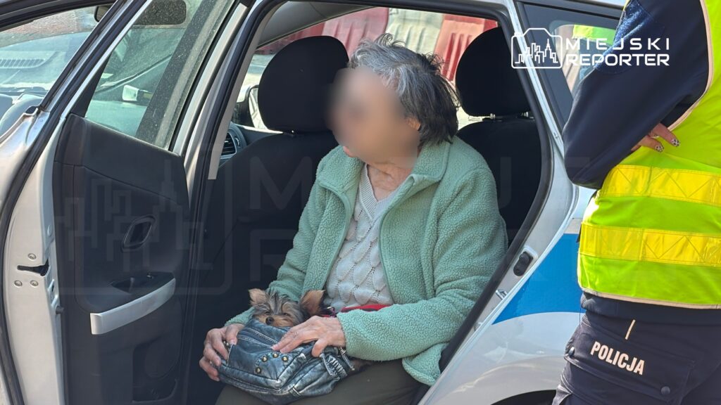 Starsza kobieta w zielonej kurtce siedzi na tylnym siedzeniu samochodu, trzymając małego psa w torebce, obok stoi policjant.