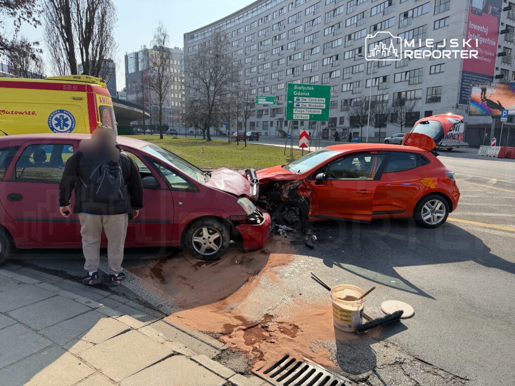 Czerwony samochód osobowy z uszkodzoną maską zderzył się z różowym pojazdem na ruchliwej ulicy, obok stoi mężczyzna.