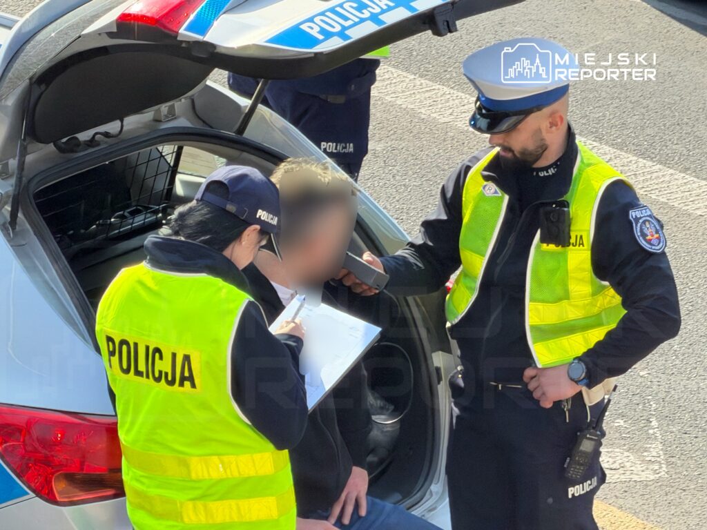 Funkcjonariusze policji przeprowadzają kontrolę drogową, rozmawiając z kierowcą przy otwartym bagażniku radiowozu.