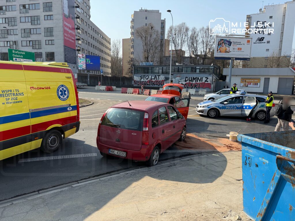 W miejscu zdarzenia drogowego, policjanci rozmawiają z kierowcą czerwoną osobówką, obok stoi karetka.