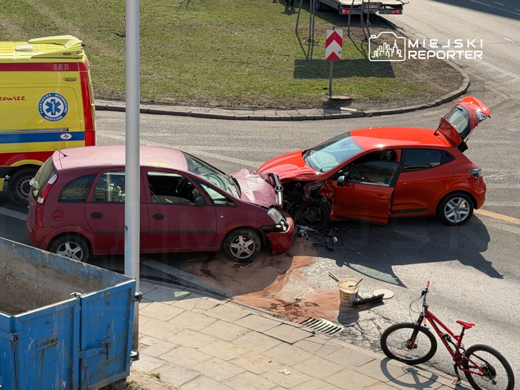 Czerwony samochód osobowy z uszkodzoną przednią częścią zderza się z różowym autem na skrzyżowaniu, obok stoi karetka.
