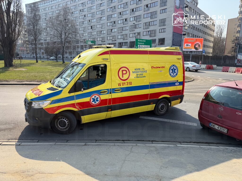 Karetka pogotowia w żółto-czerwonych barwach zatrzymana na skrzyżowaniu w miejskim otoczeniu.