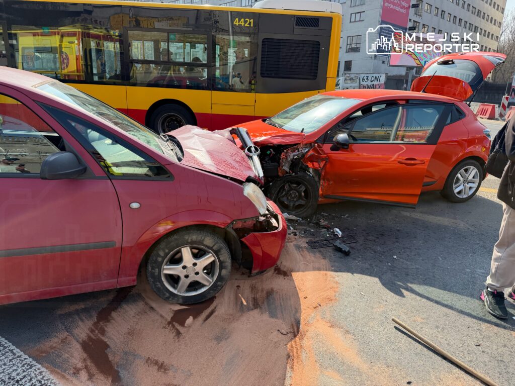 Dwa zderzone samochody osobowe na ulicy, jeden czerwony z uszkodzeniem przodu, obok mężczyzna w jasnych spodniach.
