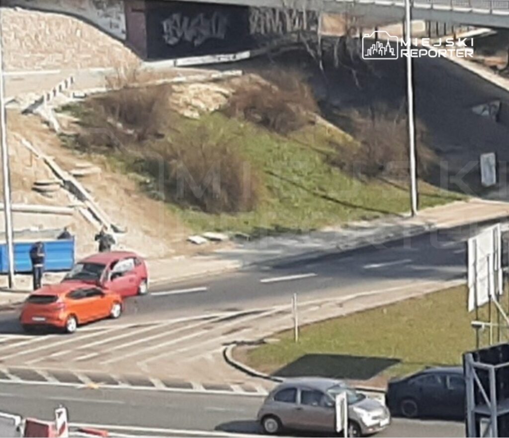 Dwa samochody zatrzymane na skrzyżowaniu, obok stoi grupa ludzi, a w tle widoczny most i zieleń.