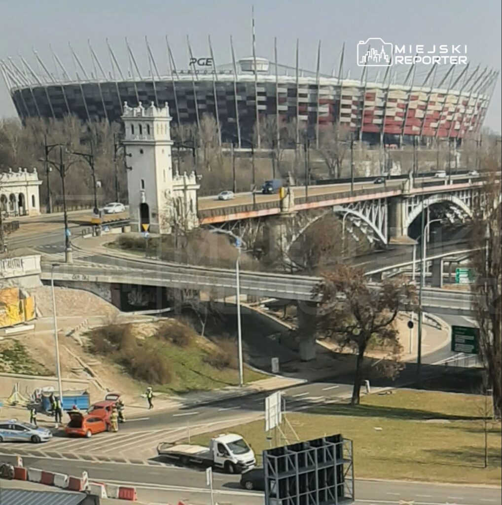Na pierwszym planie widać robotników przy czerwonym samochodzie na skrzyżowaniu, a w tle nowoczesny stadion z charakterystycznymi iglicami.
