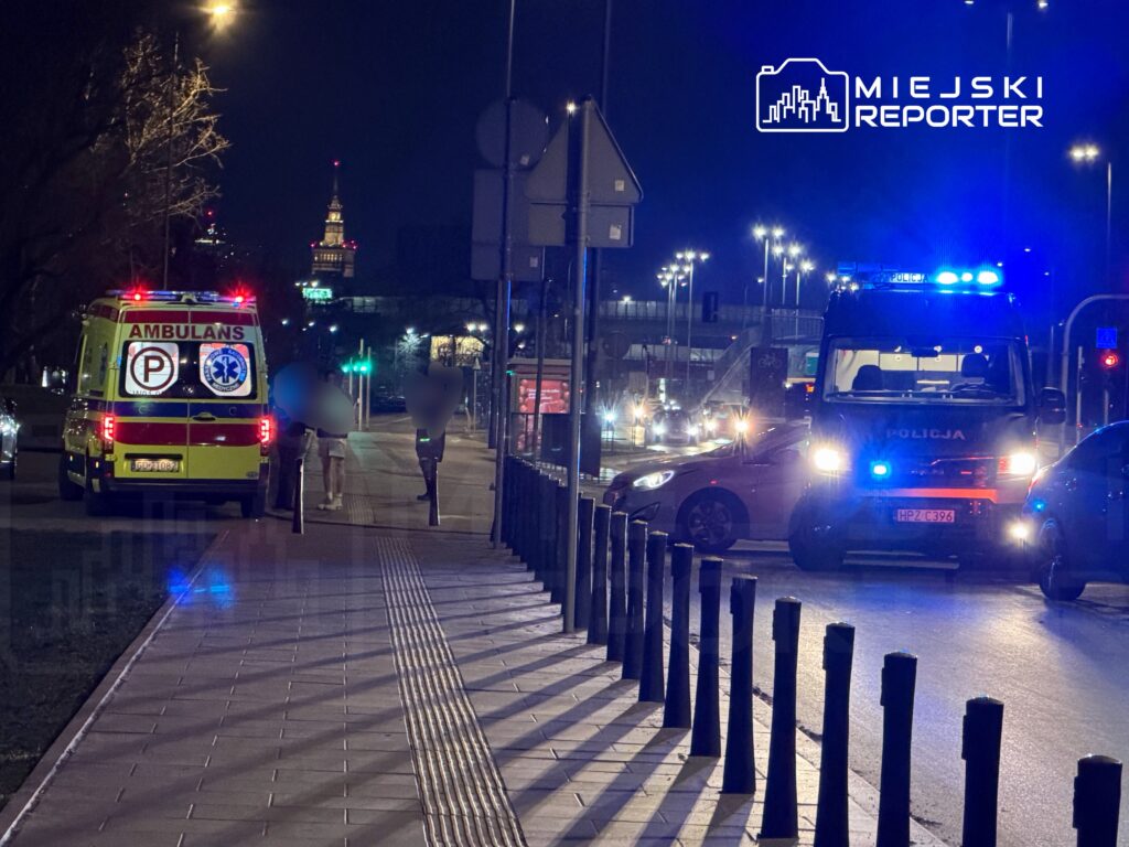 Ambulans z włączonymi sygnałami świetlnymi zatrzymany na chodniku, obok policja i piesi na oświetlonej ulicy w nocy.