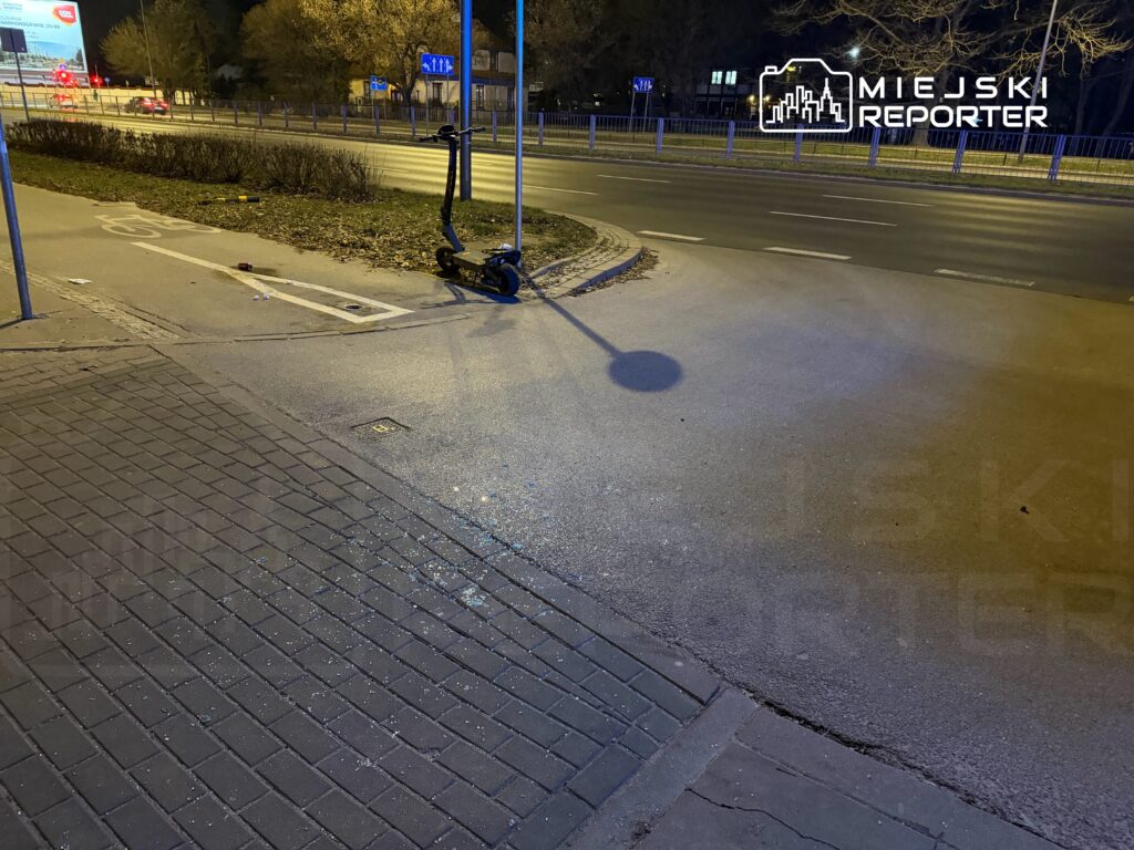 Na chodniku przy drodze stoi czarna hulajnoga elektryczna, obok widać fragmenty szkła i liście.