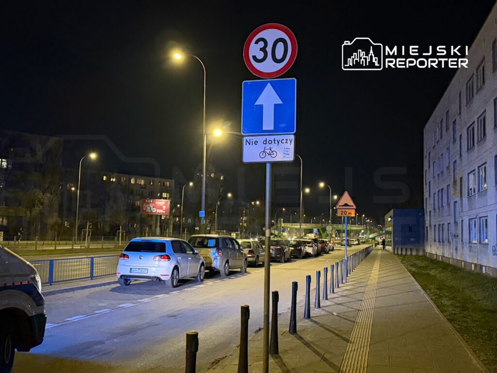 Ruchliwa ulica nocą z samochodami zaparkowanymi wzdłuż chodnika, oznakowanie drogowe z limitem prędkości 30 km/h.