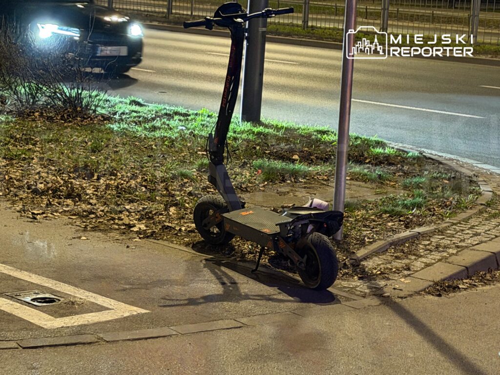 Zielona elektryczna hulajnoga zaparkowana obok latarni na trawniku przy ruchliwej ulicy nocą.