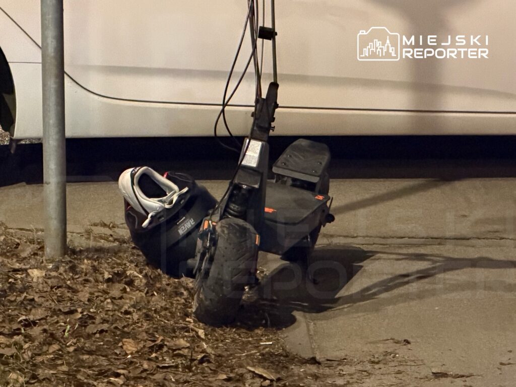 Skuter elektryczny oparty na słupku, obok znajduje się kask i plecak, w tle biały samochód.