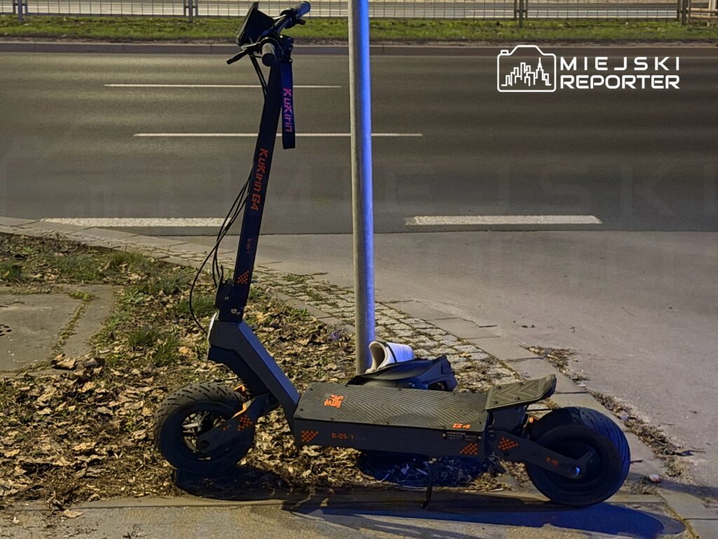 Na chodniku stoi elektryczna hulajnoga z widoczną marką, obok latarni, otoczona opadłymi liśćmi.