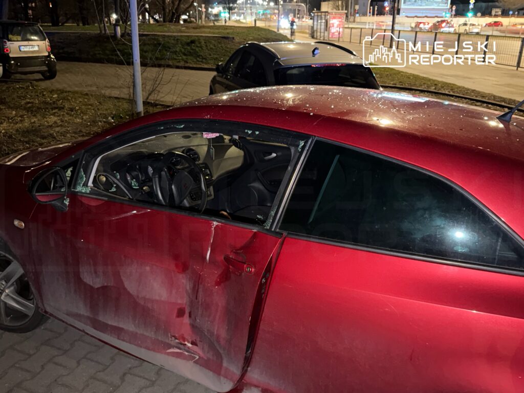 Czerwony samochód z wybitymi szybami i uszkodzoną karoserią stoi na parkingu nocą.