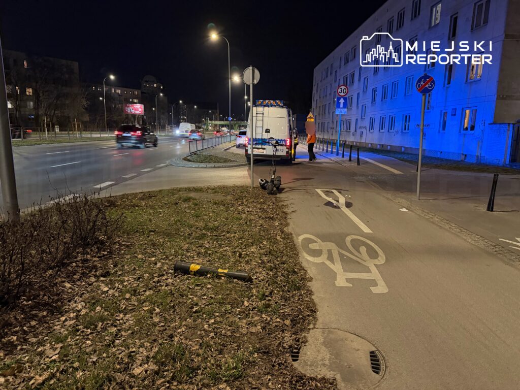 Na poboczu drogi stoi policyjny samochód, a funkcjonariusz rozmawia z osobą w pomarańczowej kamizelce. W tle widać ruch uliczny.