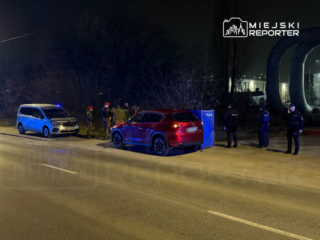 Grupa policjantów w mundurach prowadzi czynności na poboczu drogi, obok zaparkowanych pojazdów służbowych.