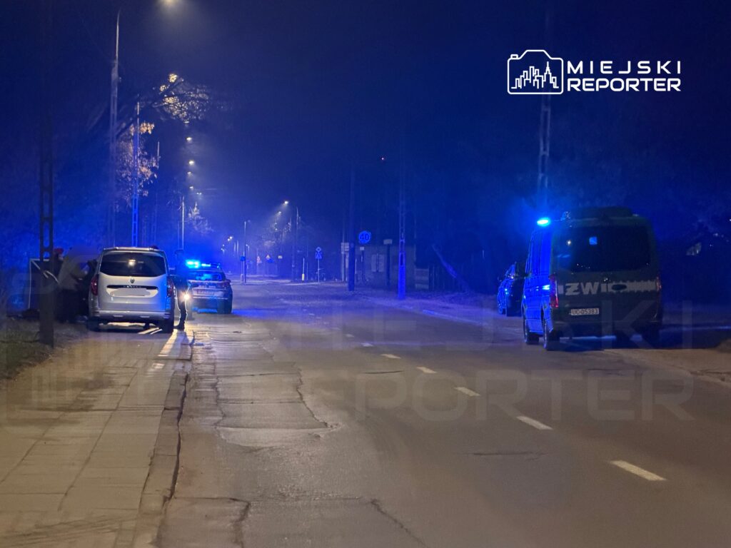 Nocna ulica oświetlona niebieskimi światłami radiowozów policji i pojazdu specjalistycznego, zaparkowane samochody.