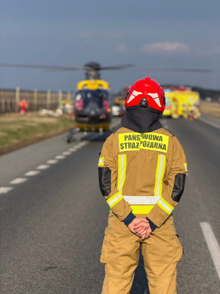Strażak w hełmie obserwujący lądowanie śmigłowca ratunkowego na drodze, w tle pojazdy służb ratunkowych.