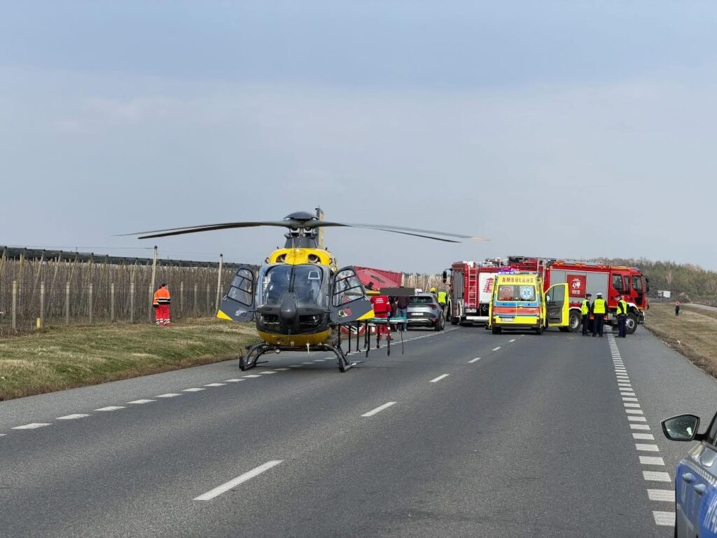 Śmigłowiec ratunkowy ląduje na drodze, obok stoją pojazdy służb ratunkowych oraz ubrani w kamizelki odblaskowe ratownicy.
