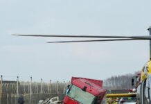 Czerwony ciężarowy pojazd leży na boku przy drodze, obok interweniujący strażacy i helikopter ratunkowy.