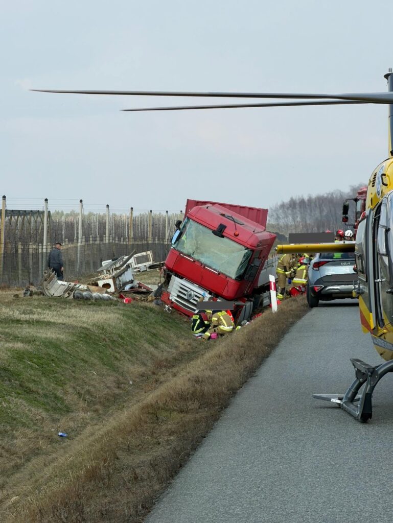 Czerwony ciężarowy pojazd leży na boku przy drodze, obok interweniujący strażacy i helikopter ratunkowy.