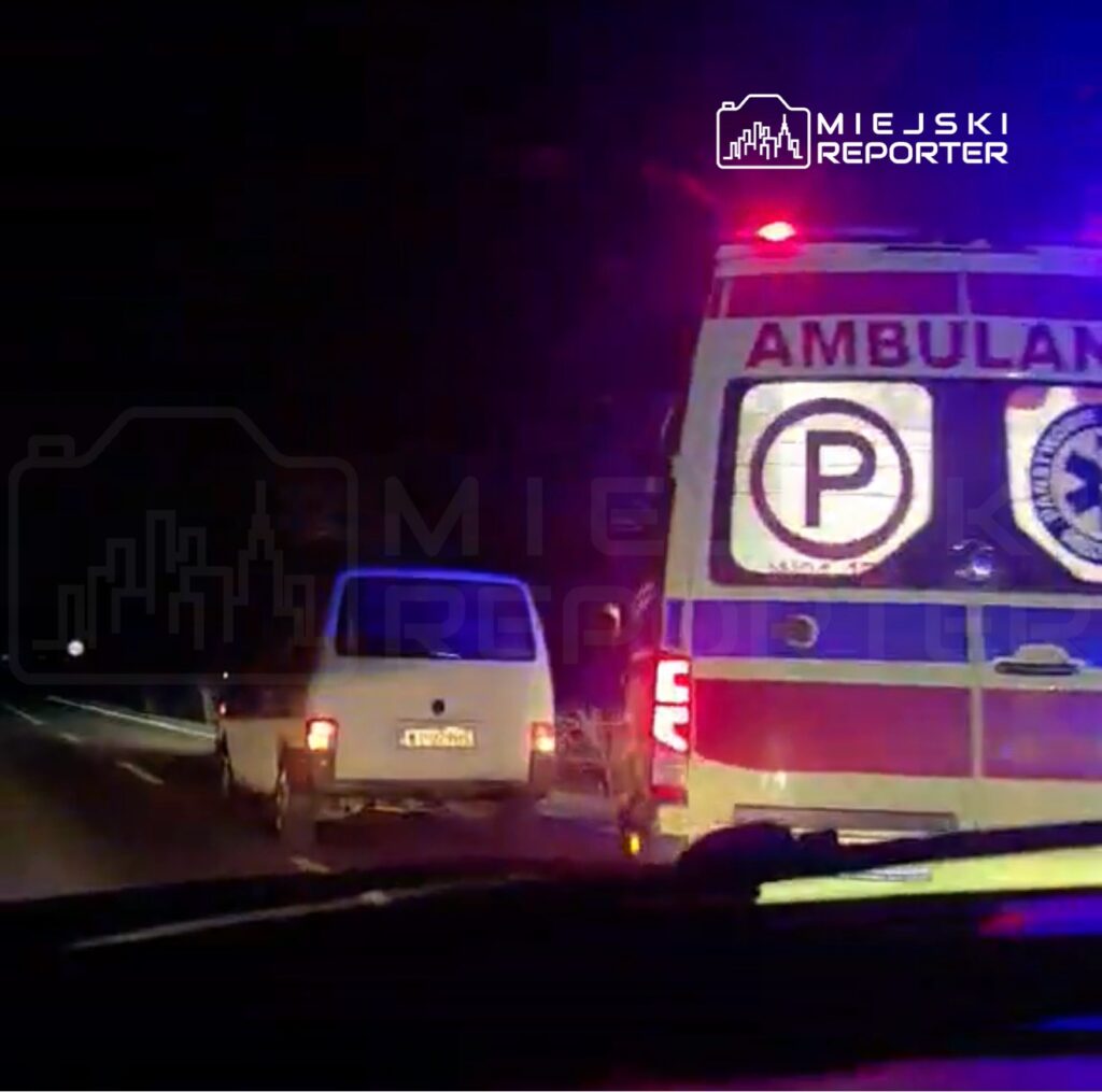 Ambulans z włączonymi sygnałami świetlnymi zatrzymany na drodze obok białego pojazdu dostawczego w nocy.