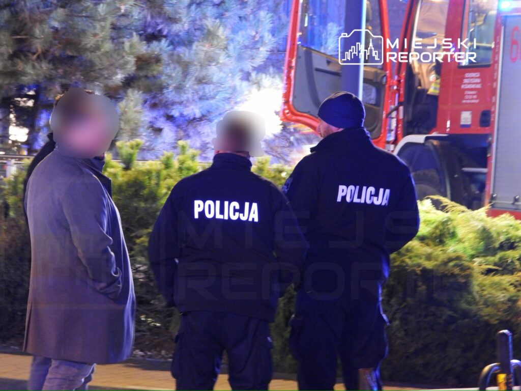 Mężczyzna w szarym płaszczu rozmawia z dwoma policjantami w mundurach, w tle widać pojazd strażacki.