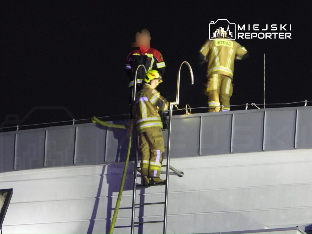 Strażacy w mundurach przystępują do akcji ratunkowej na dachu budynku w nocy, używając drabiny i sprzętu.