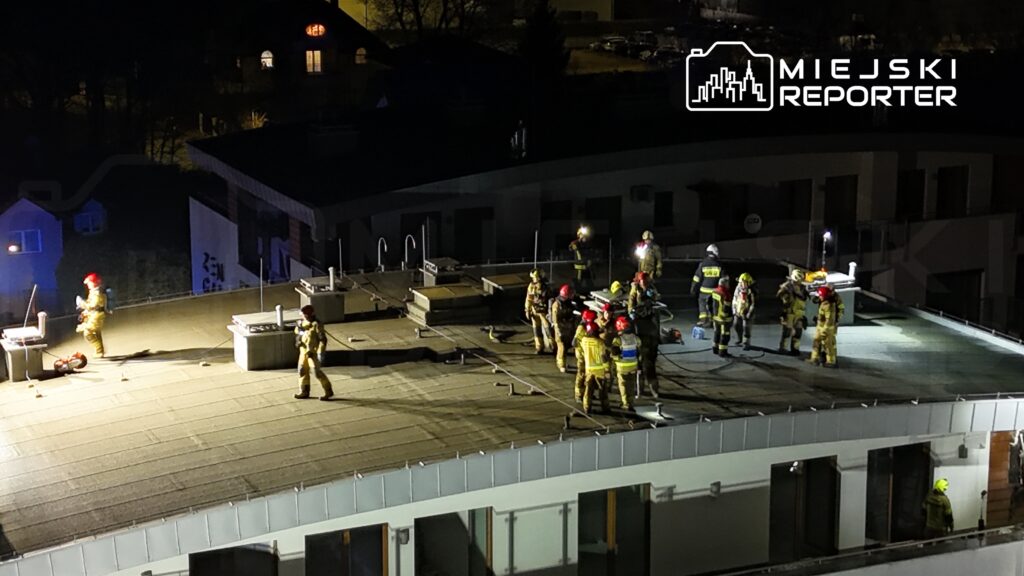 Grupa strażaków w strojach ochronnych prowadzi działania na dachu budynku w nocy, używając latarek.