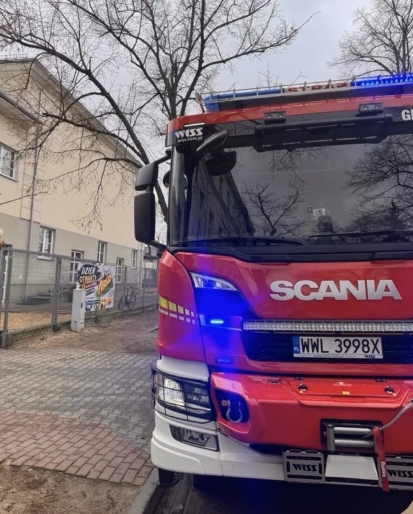 Czerwony wóz strażacki marki Scania z włączonymi sygnalizacjami świetlnymi stoi na ulicy obok budynku.