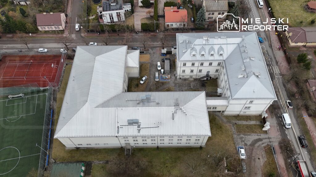 Widok z lotu ptaka na kompleks budynków o jasnych fasadach z metalowymi dachami, w otoczeniu boisk sportowych i ulic.