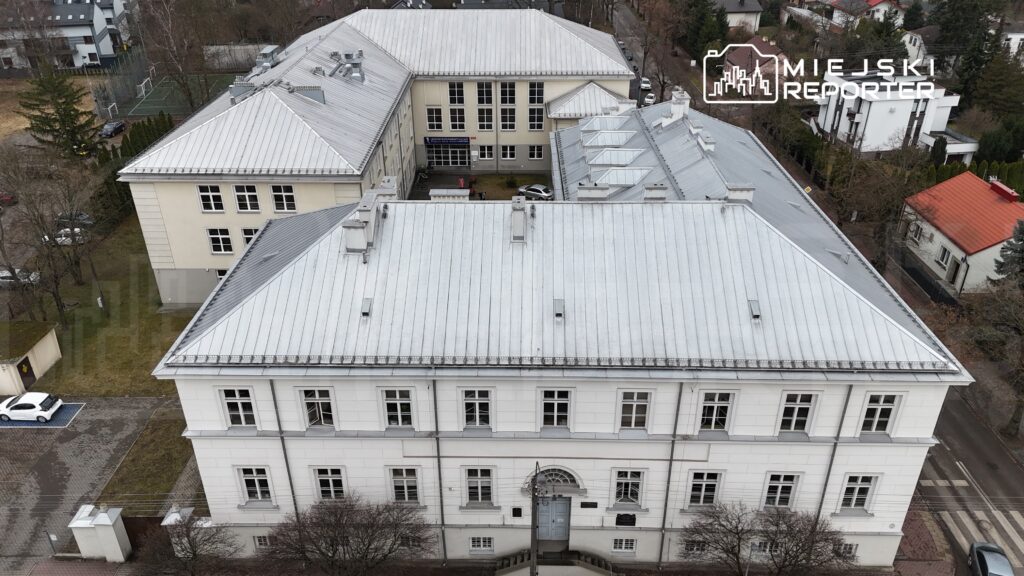 Budynek z białą elewacją i metalowym dachem otoczony zielenią oraz parkingiem, widok z góry.
