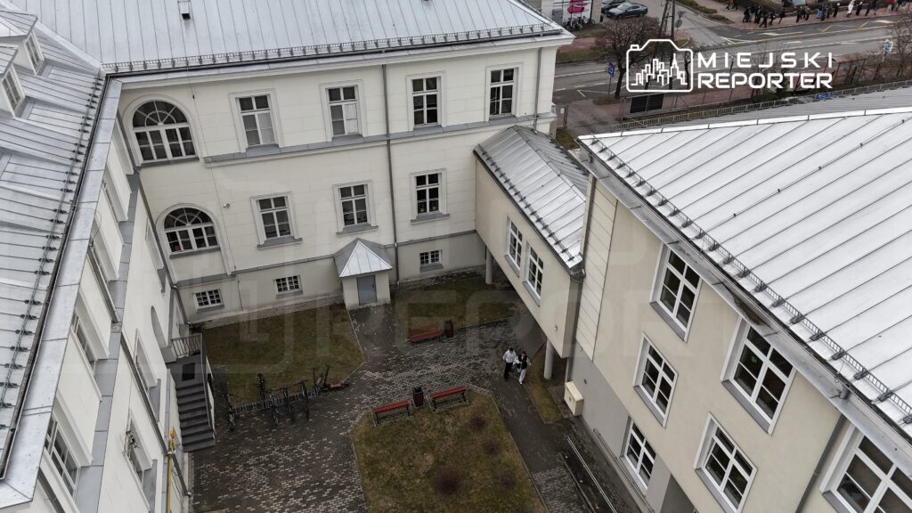 Dwóch ludzi spaceruje po dziedzińcu otoczonym białymi budynkami z metalowymi dachami.