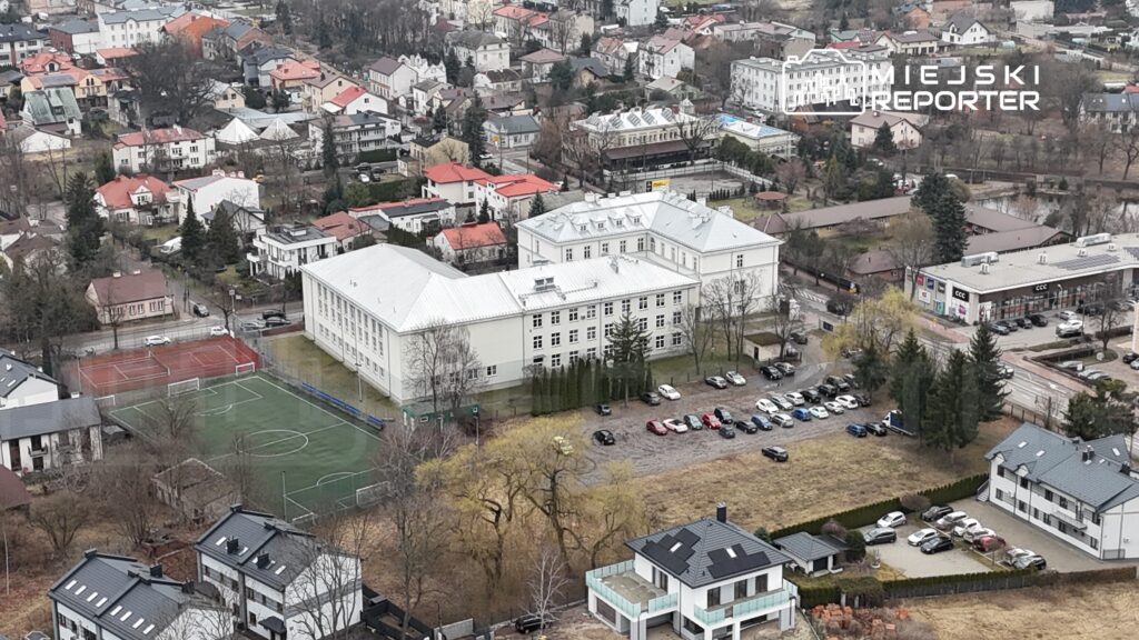 Widok na nowoczesny budynek szkoły otoczony boiskami sportowymi i parkingiem w zabudowanej okolicy.