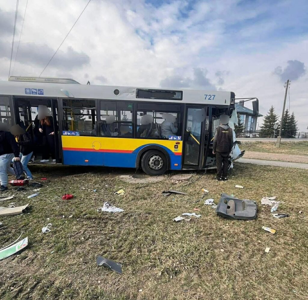 Zniszczony autobus komunikacji miejskiej z otwartymi drzwiami, obok grupka osób oraz porozrzucane fragmenty pojazdu na trawie.