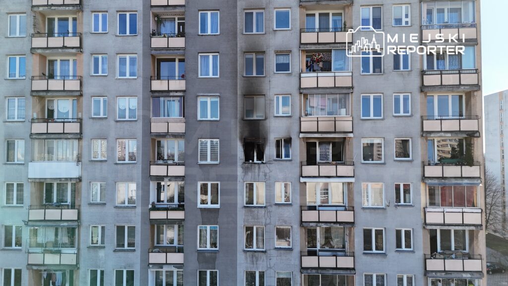 Na elewacji bloku widać ślady po pożarze, czarny dym wokół okna na drugim piętrze. Sąsiednie balkony z roślinami.