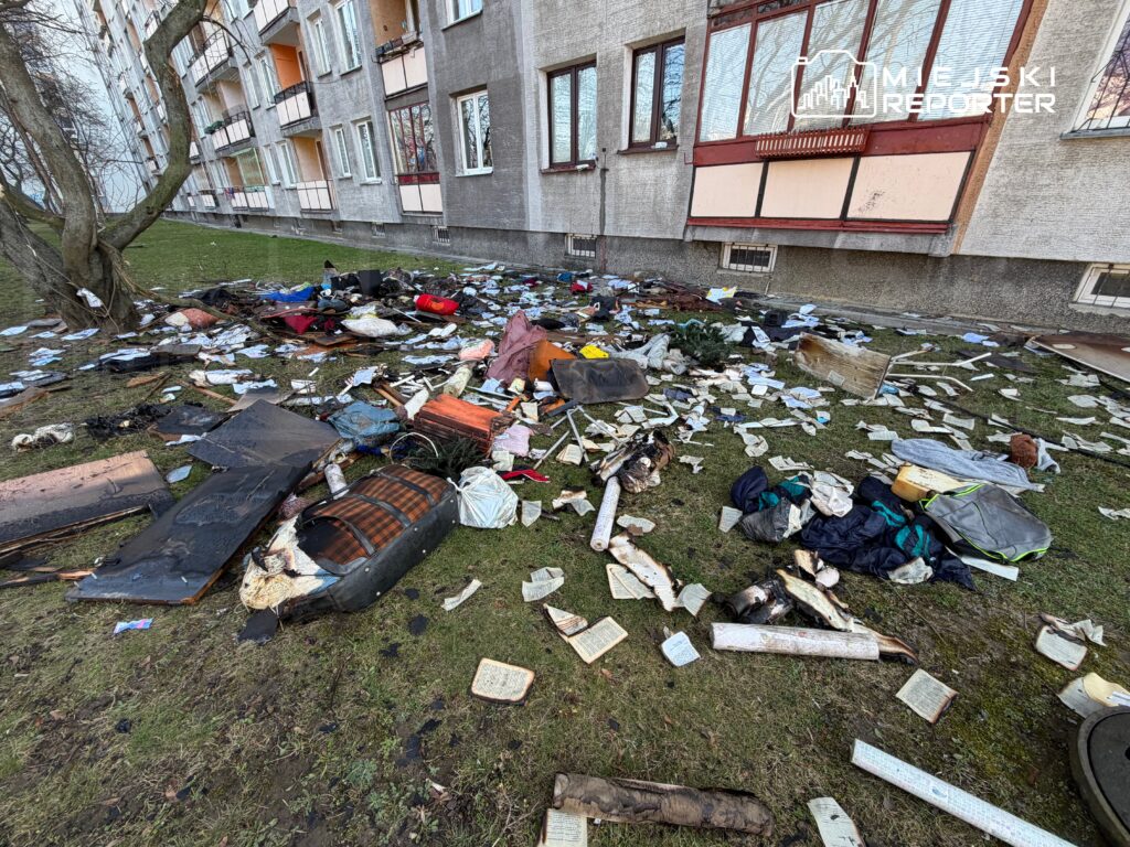 Na trawie przed budynkiem mieszkalnym leży stos odpadów, w tym zniszczone meble, ubrania i porozrzucane kartki papieru.