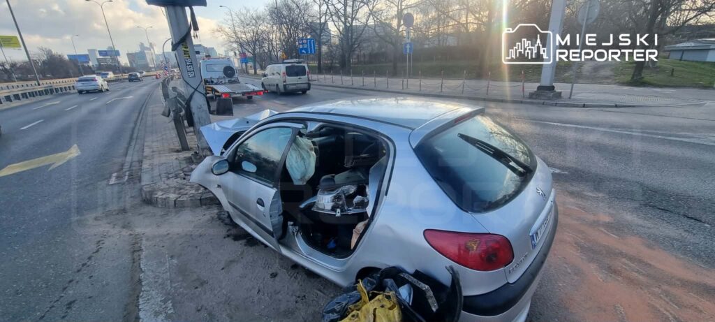 Uszkodzony samochód osobowy zaparkowany przy krawężniku, obok słupa drogowego, na ruchliwej ulicy.