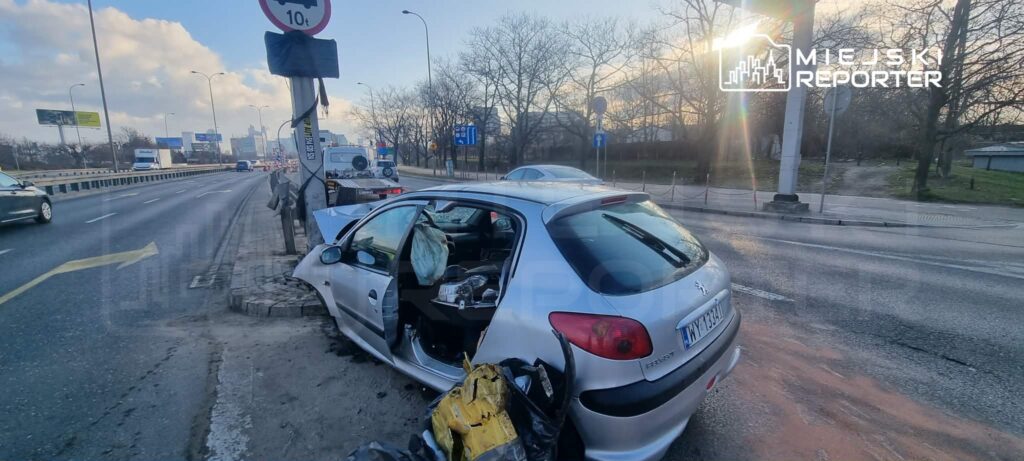 Uszkodzony samochód osobowy stoi przy drodze, z otwartymi drzwiami i uszkodzoną karoserią, obok znak drogowy.