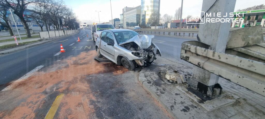 Rozbity samochód osobowy stoi na poboczu drogi, obok ustawione pachołki ostrzegawcze. W tle widać nowoczesne budynki biurowe.