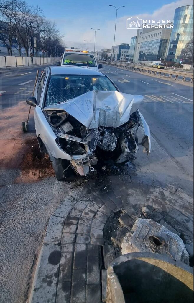 Zniszczony samochód osobowy z wgniecionym przodem stoi na poboczu ulicy, obok widoczny wóz policyjny.