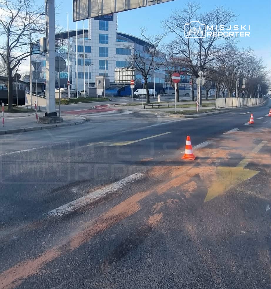 Droga z widocznymi znakami drogowymi, pachołkami i zarysami farby na jezdni w otoczeniu nowoczesnego biurowca.