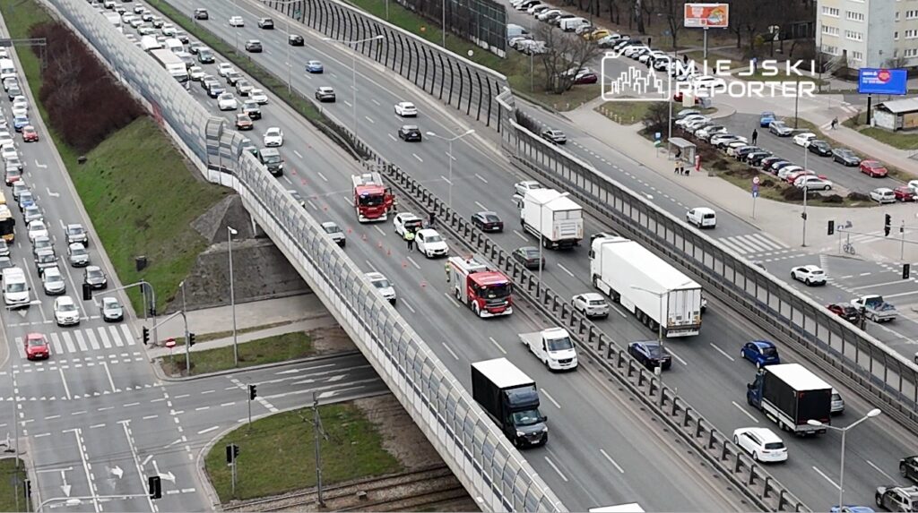 Ruch samochodowy na drodze, z widocznymi pojazdami ratunkowymi oraz korkiem w obie strony.