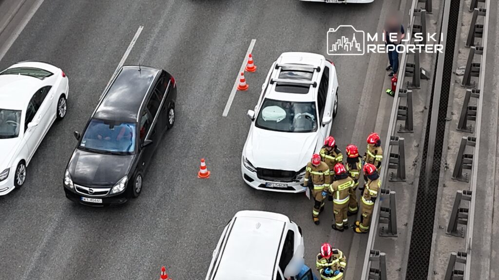 Grupa strażaków w hełmach omawia sytuację obok białego SUV-a na ruchliwej drodze, otoczonego przez samochody.