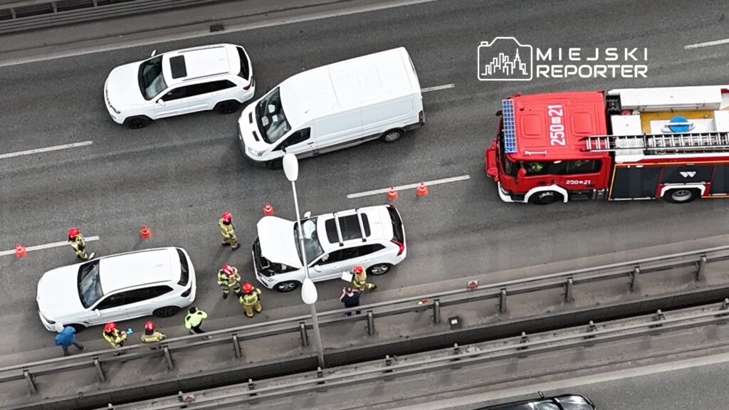 Grupa strażaków w kaskach bada uszkodzone samochody na ruchliwej drodze, obok stoi wóz strażacki.