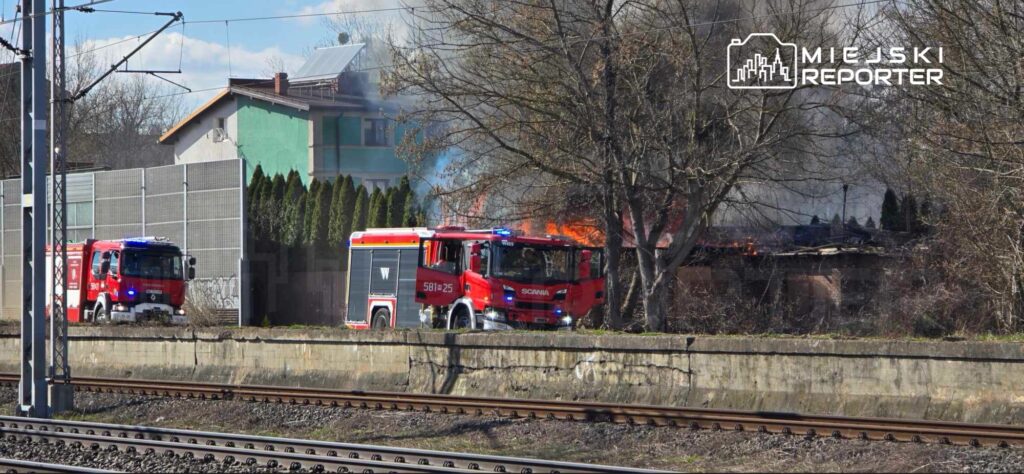 Czerwony wóz strażacki z włączonymi sygnałami przejeżdża obok palącego się budynku w otoczeniu drzew.