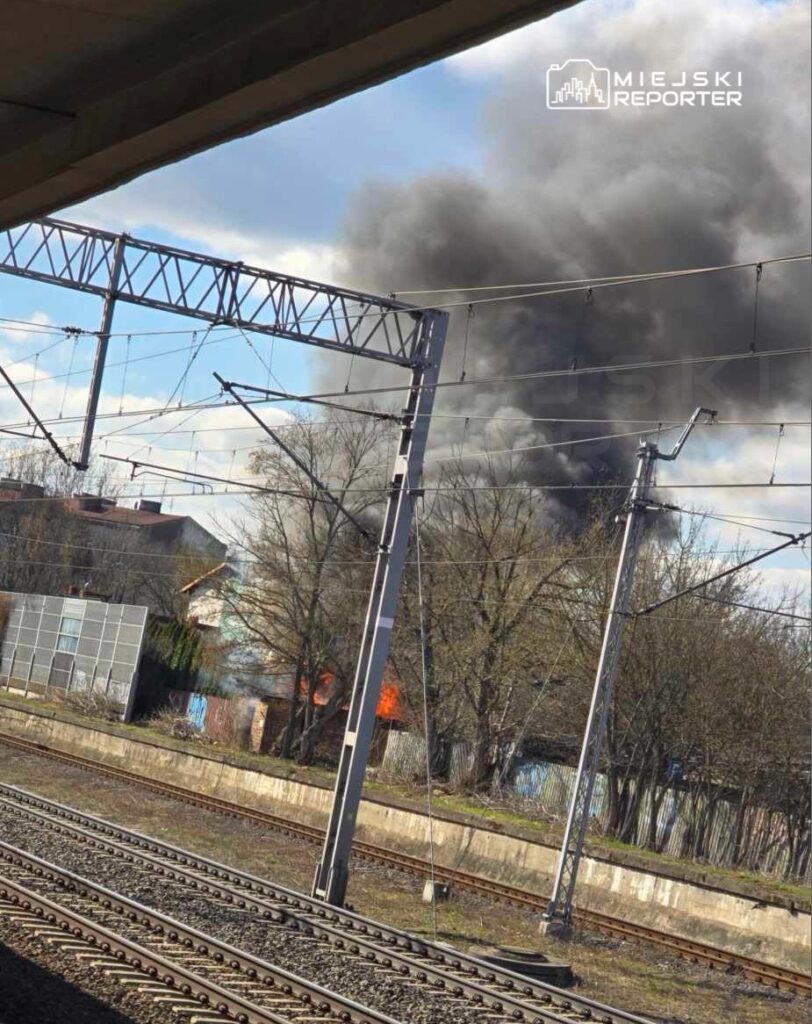 Czarny dym unoszący się z płonącego budynku w tle, widok z peronu kolejowego.